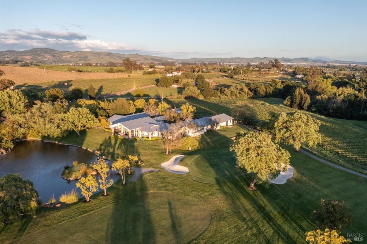 Aerial view of a large estate surrounded by vineyards and rolling hills. The property features a pond with a small island, a main house with a large lawn, and a golf putting area with sand traps. The surrounding landscape includes trees, manicured gardens, and a mix of open fields and dense greenery.