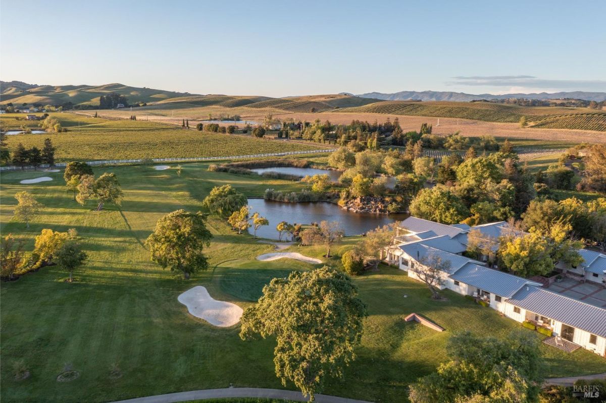 Aerial view of a property surrounded by vineyards and rolling hills. The landscape includes ponds, green fields, and sand traps, suggesting a golf course area. A large modern building complex with metal roofing is situated near the water, with trees and pathways surrounding it.