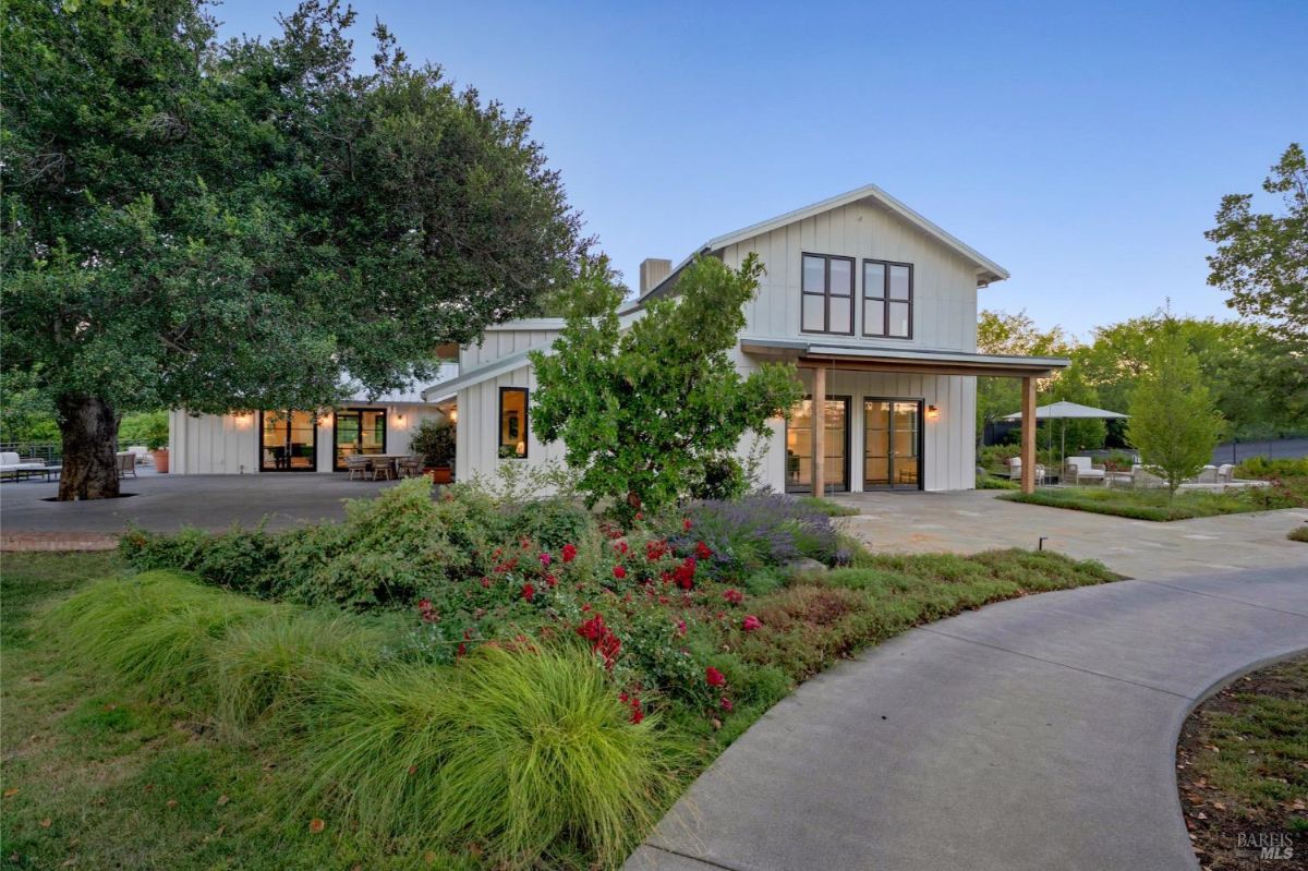 A two-story white modern farmhouse features large windows, a covered front patio, and landscaping with a mix of shrubs and flowers. A paved pathway curves through the lawn, leading to the house. Outdoor seating areas are visible to the right, surrounded by greenery and small trees.