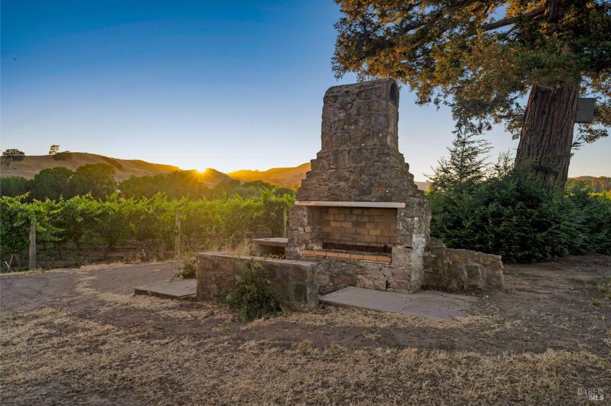 An outdoor stone fireplace with a rectangular opening and a weathered stone structure. It is set on a patch of bare ground with a vineyard in the background. The sun is setting behind rolling hills, and a large tree with branches partially frames the right side of the image.