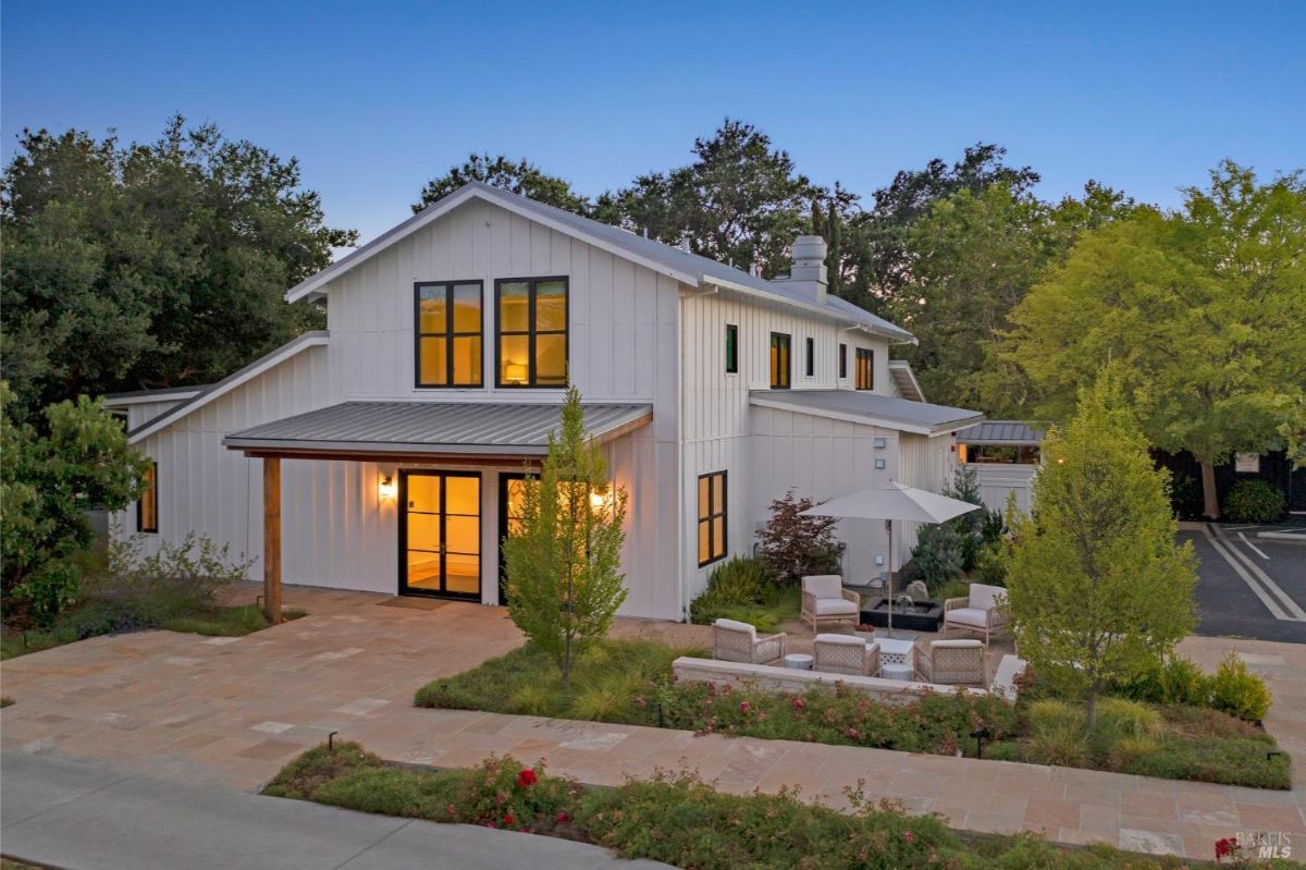 A modern white house with black-framed windows and a covered front entrance. A patio area to the right features outdoor seating under a white umbrella, surrounded by landscaped greenery. A paved walkway and well-maintained lawn extend in front of the house, with trees and shrubs in the background.