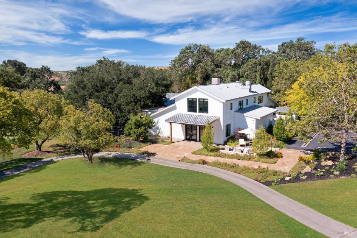 A modern two-story white building with a metal roof surrounded by landscaped greenery. A curved pathway leads up to the house, with a manicured lawn and several trees nearby. There is an outdoor patio area with seating, and the background includes dense trees and open sky.