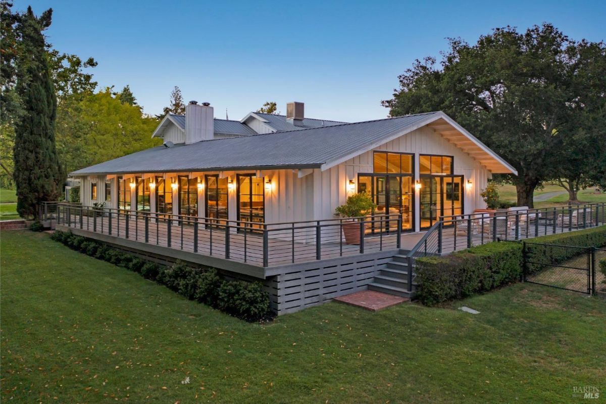  Modern single-story building with large windows and exterior lighting. A spacious wooden deck with a black metal railing wraps around the structure, providing outdoor seating space. The surrounding area features a green lawn, tall trees, and hedges, with a pathway leading up to the deck stairs. 