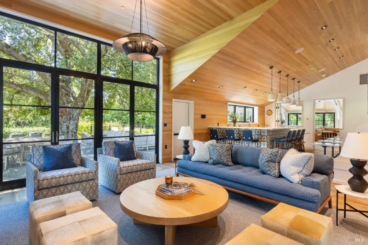  A modern living space with a wooden ceiling and large glass windows that overlook an outdoor area. The room features a blue sofa, two armchairs, two ottomans, and a round wooden coffee table in the center. In the background, a kitchen with pendant lights, a bar counter, and stools is visible.