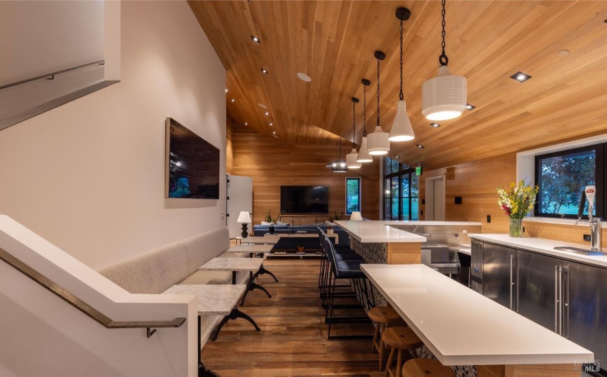 Modern interior space with a large white bar counter and high-backed stools arranged along its side. A long cushioned bench seating with small marble-topped tables and stools is positioned parallel to the bar. The wooden ceiling features pendant lights, and the space includes wood-paneled walls, a mounted TV, and a window looking out to an outdoor area.