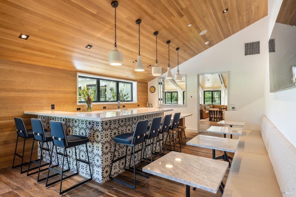 Modern interior space with a large white bar counter and high-backed stools arranged along its side. A long cushioned bench seating with small marble-topped tables and stools is positioned parallel to the bar. The wooden ceiling features pendant lights, and the space includes wood-paneled walls, a mounted TV, and a window looking out to an outdoor area.