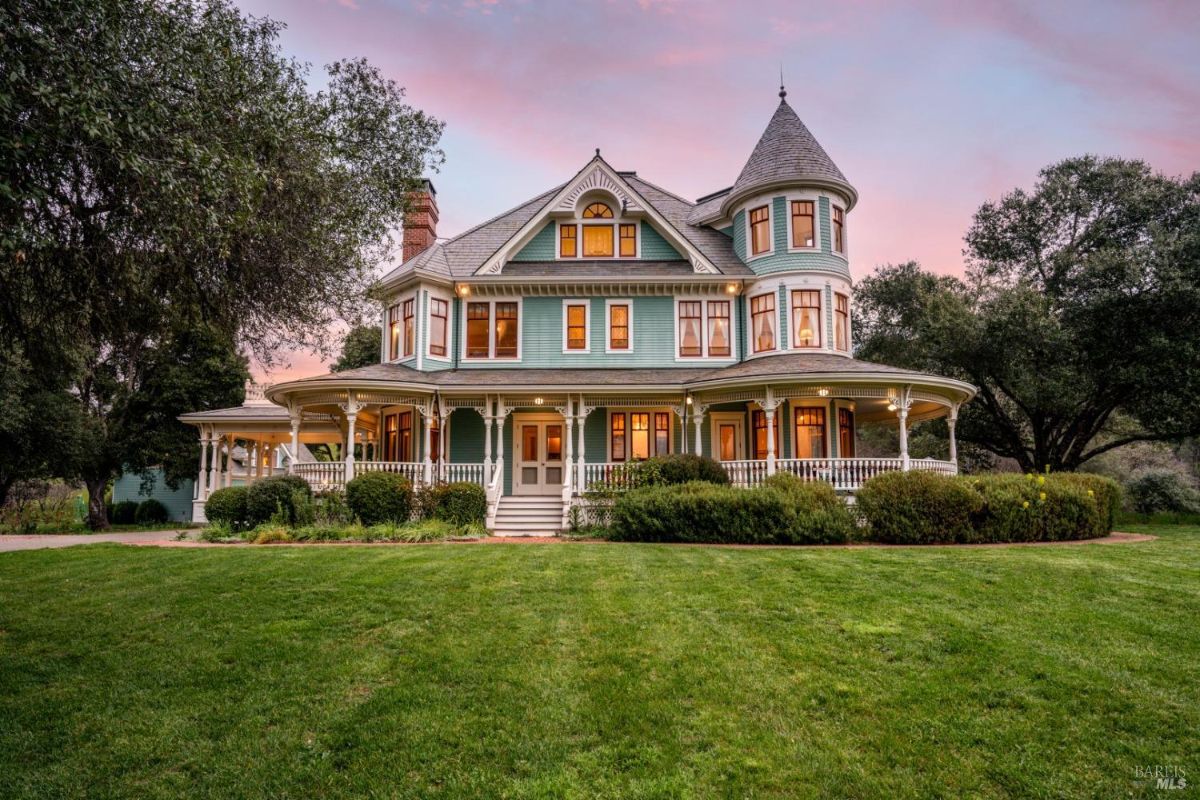 A beautifully restored Victorian-style home with a wraparound porch and vibrant green exterior under a soft sunset sky. The lush green lawn complements the intricate architectural details.