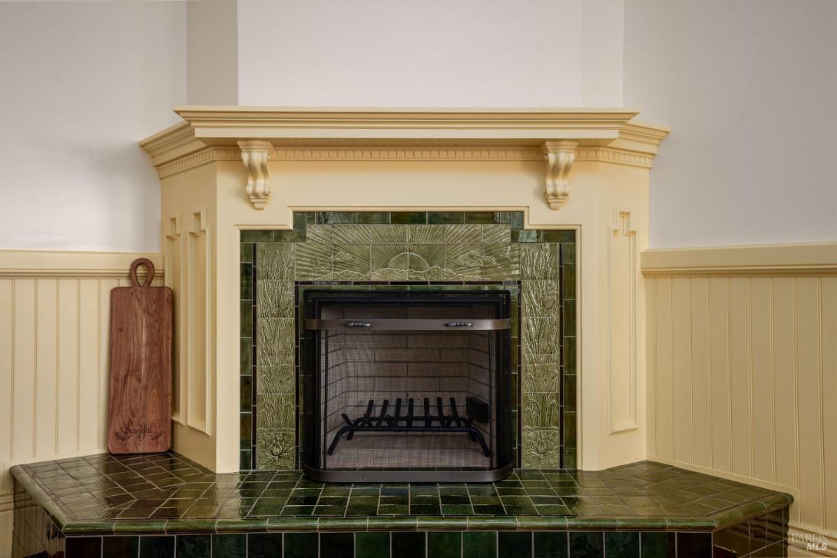 A detailed view of a beautifully designed fireplace with green ceramic tiles and intricate embossed patterns. The wooden mantle and cream-colored paneling add an elegant and cozy touch