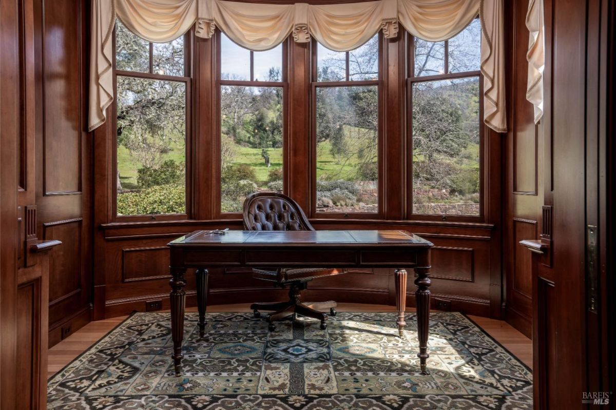 A luxurious home office with a large wooden desk and leather chair, surrounded by rich wooden paneling and a wall of windows offering picturesque views of the lush outdoors.