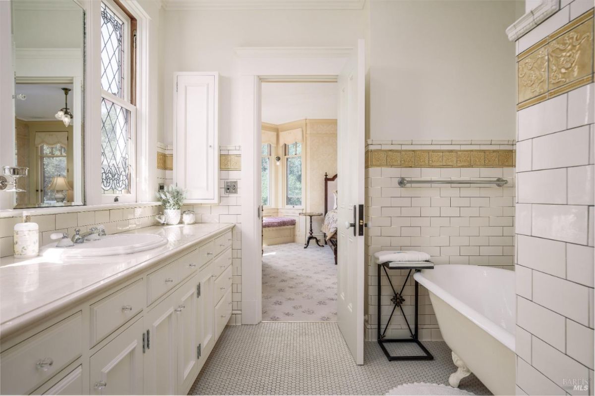 A bright bathroom featuring white subway tiles with golden accents. A clawfoot tub adds vintage charm, and the long vanity with a white countertop provides ample storage. A window brings in natural light, and the space connects to a nearby bedroom.