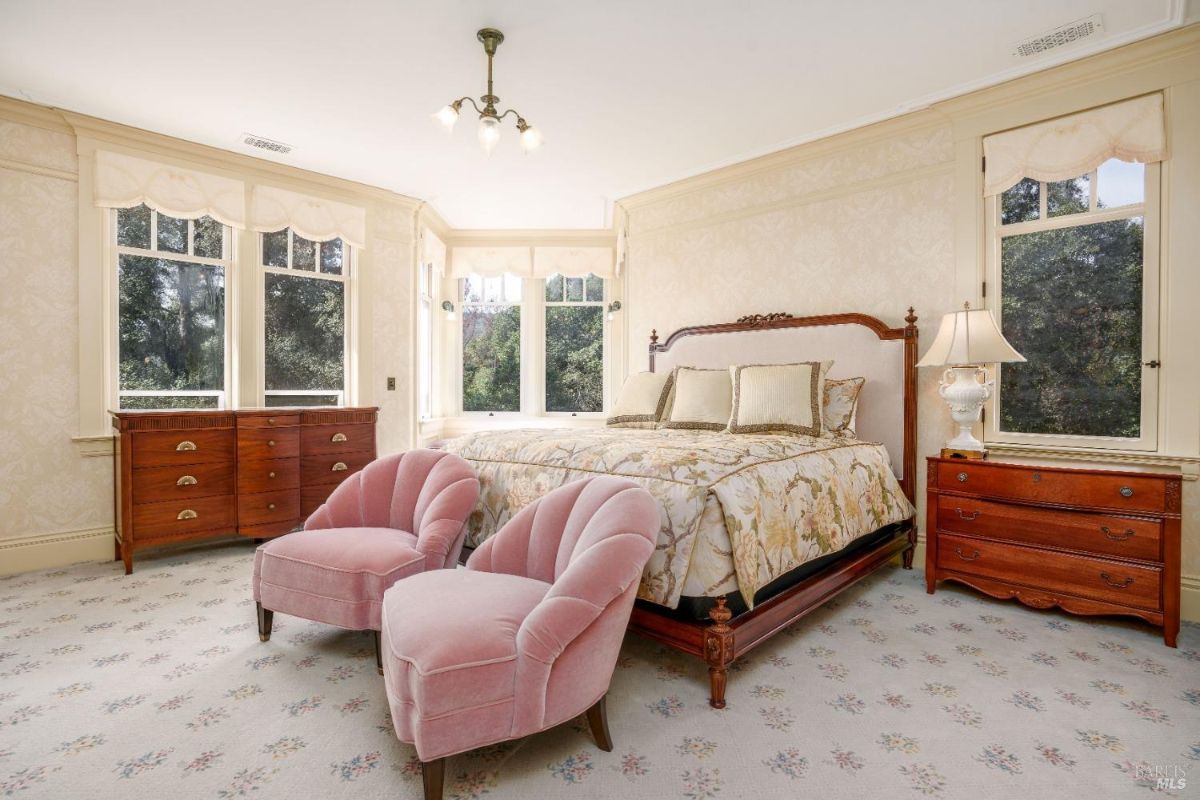 Spacious master bedroom with an elegant floral-patterned bedspread and a headboard. Two blush pink armchairs provide a touch of luxury. The room features multiple large windows framed with light curtains, filling the space with natural light.