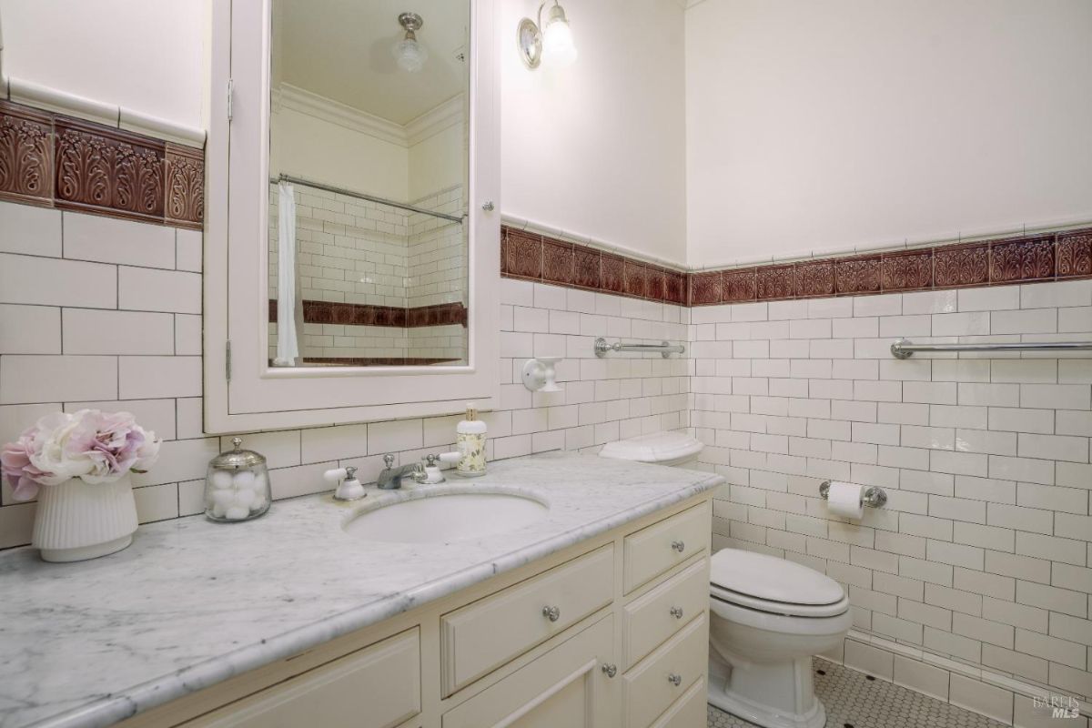 Bathroom with white subway tiles and burgundy accent tiles adding texture. It features a marble countertop, a simple yet elegant sink, and a vintage-inspired mirror. A tiled bathtub is partially visible, enhancing the classic aesthetic.