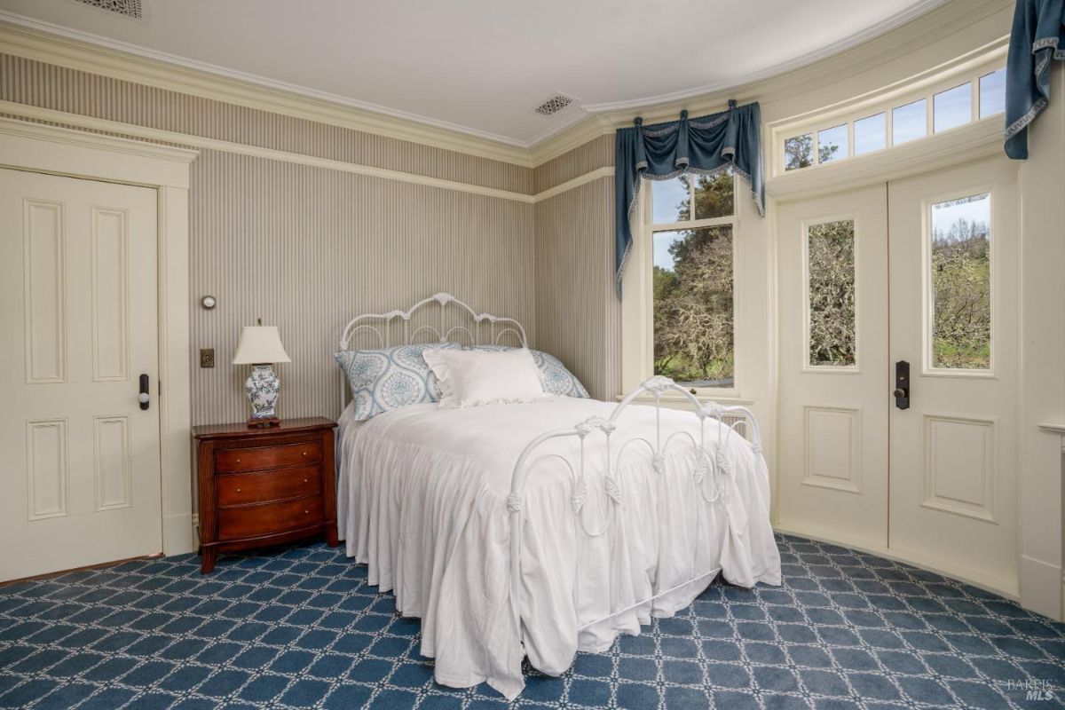 Bedroom featuring a white metal frame bed with elegant white bedding. The room has soft gray patterned wallpaper, blue curtains, and carpet with a diamond pattern. French doors with window panels add brightness and connect the room to the outdoors.