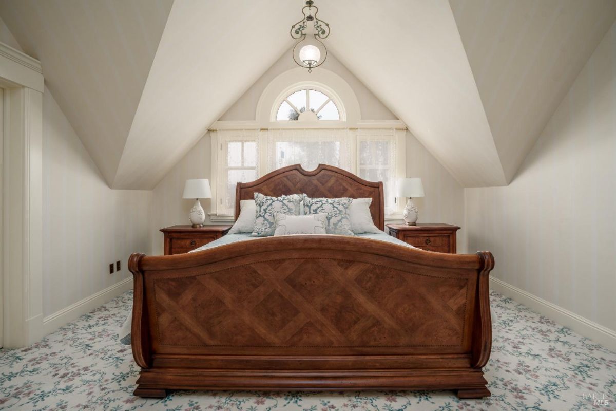 A unique bedroom with a sloped ceiling and an arched window above the bed. The wooden bed frame has a herringbone design, complemented by matching nightstands. The cozy space has floral carpeting and soft cream-colored walls.