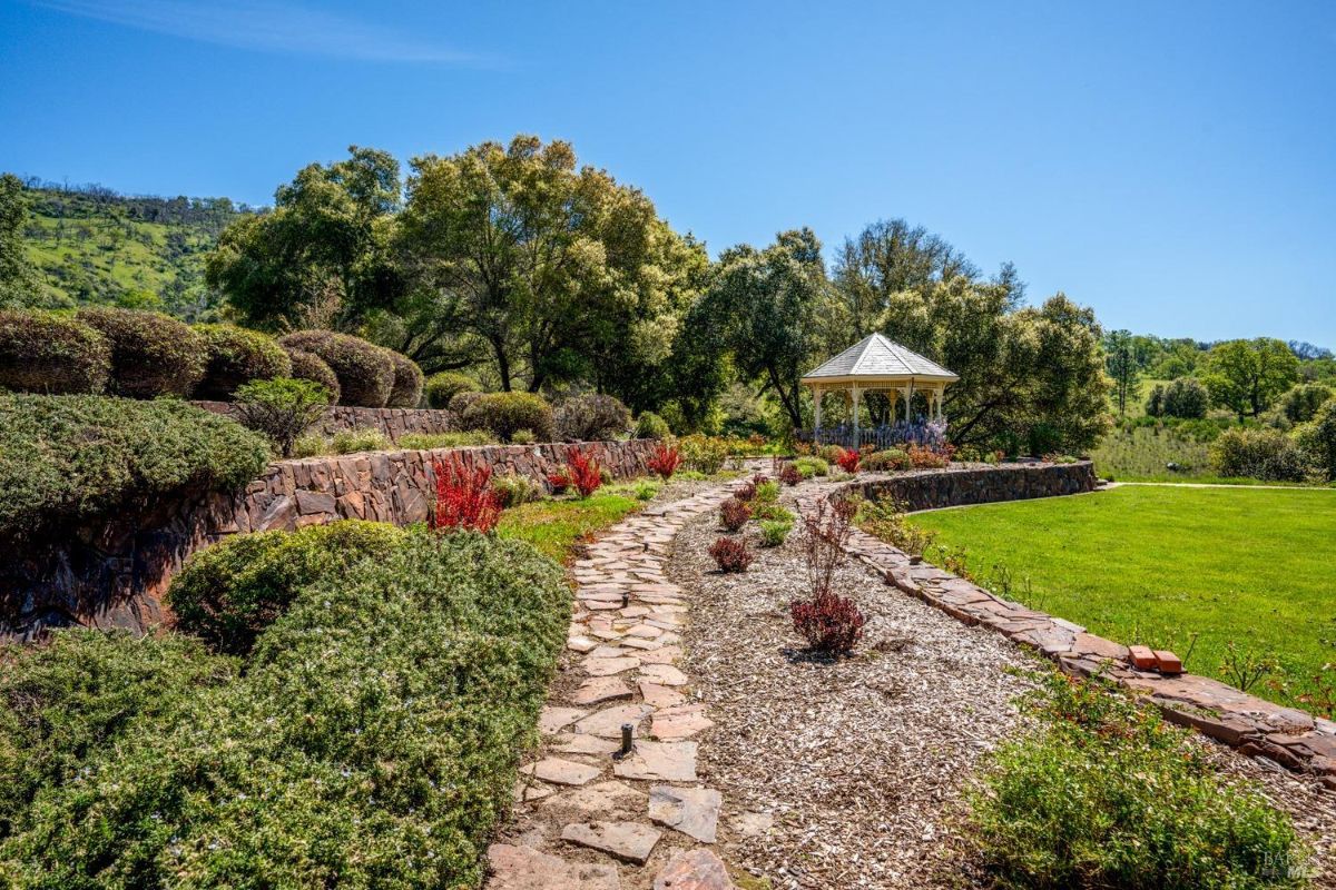  A stone pathway meandering through landscaped gardens with colorful shrubs and flowers, leading to a gazebo. The elevated greenery and stone wall create a tranquil environment.