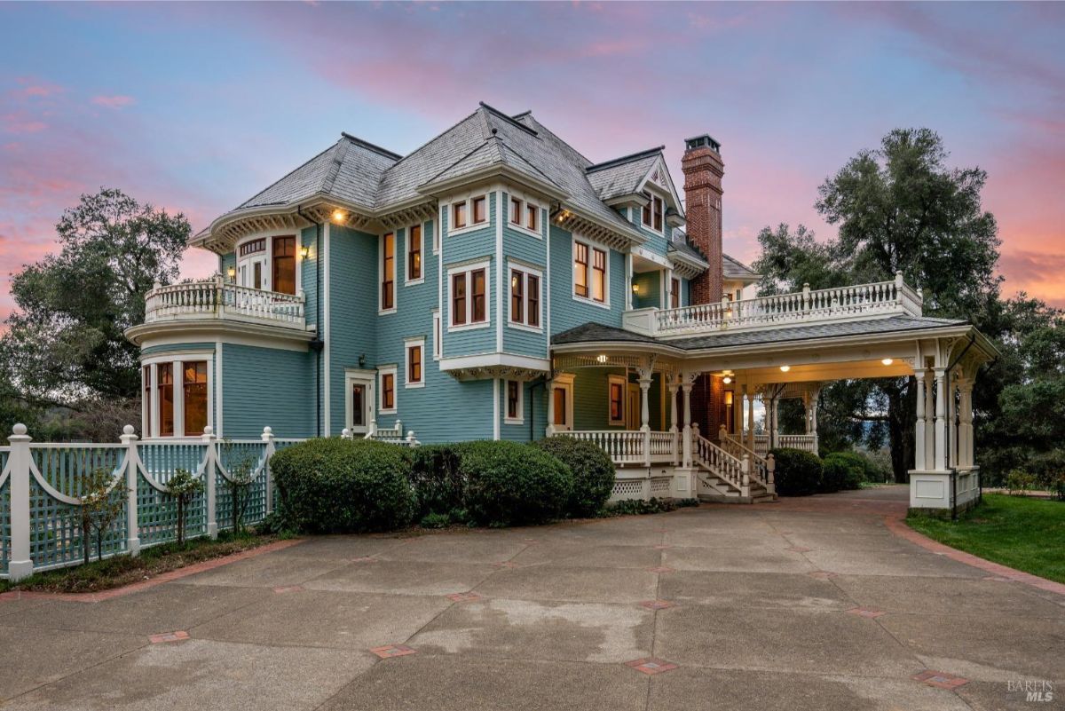 The side view of the same Victorian home showcasing its elegant bay windows, a second-story balcony, and a covered driveway entry. The paved driveway adds to the grand entrance.