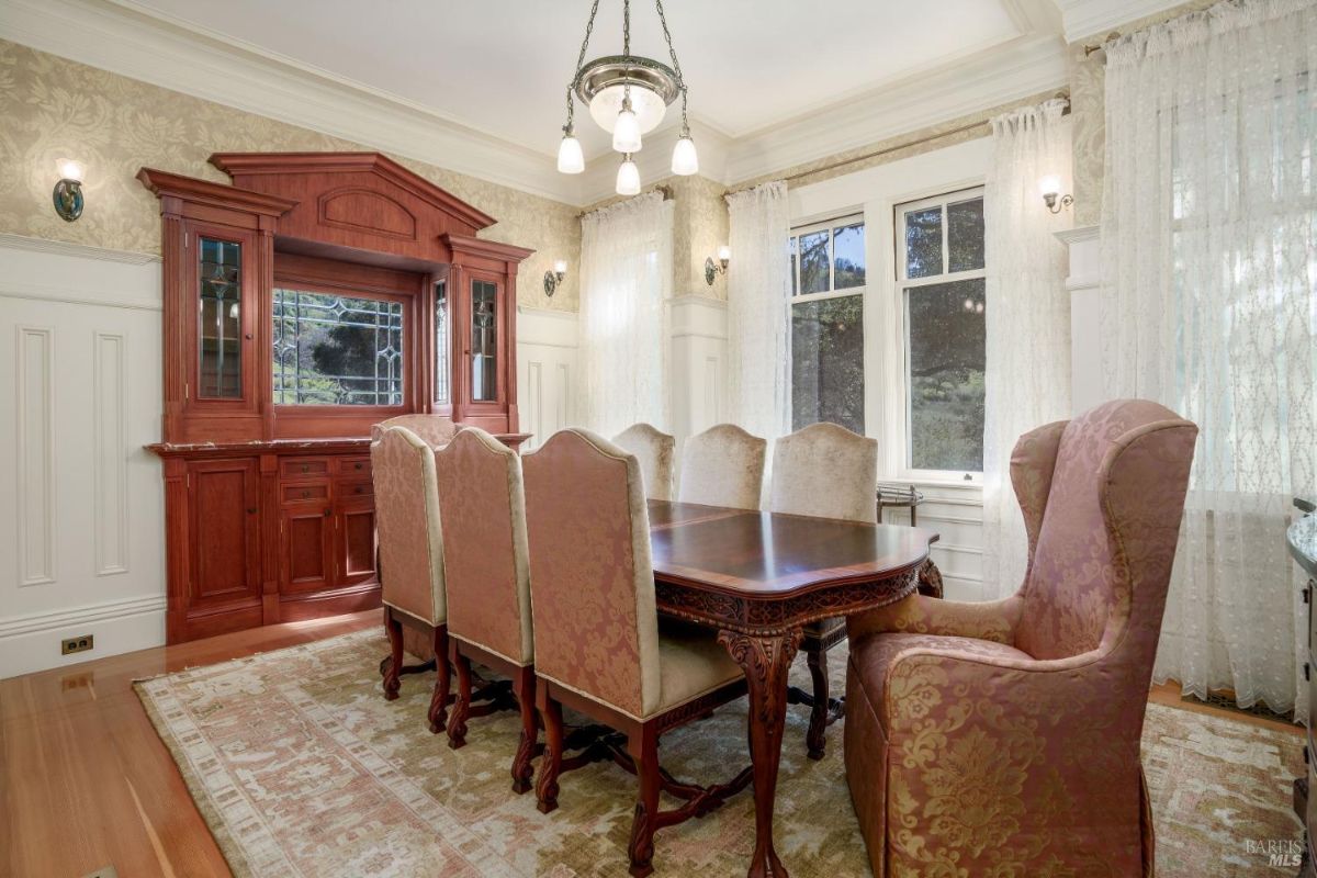 Dining table with eight upholstered chairs. The centerpiece is a built-in wooden hutch with intricate stained glass and decorative cabinetry. The ambiance is warm, enhanced by light curtains and traditional wainscoting.