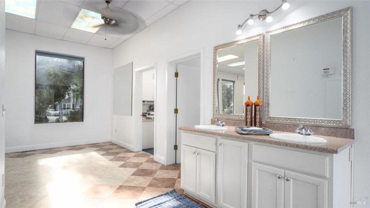 A spacious and brightly lit bathroom featuring a double vanity with two mirrors, ample counter space, and white cabinetry. Large windows allow natural light to flood the room.