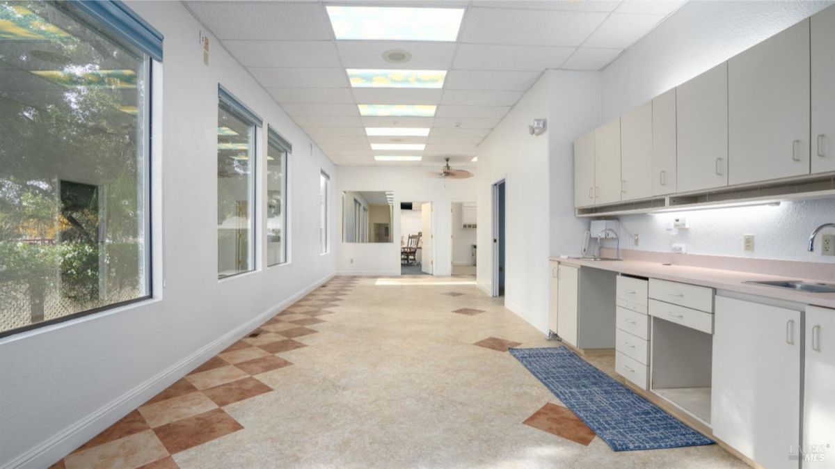 A long, sunlit hallway with large windows overlooking the outdoors, checkered flooring, and built-in cabinetry. This space appears multifunctional, with a practical layout suitable for various uses.