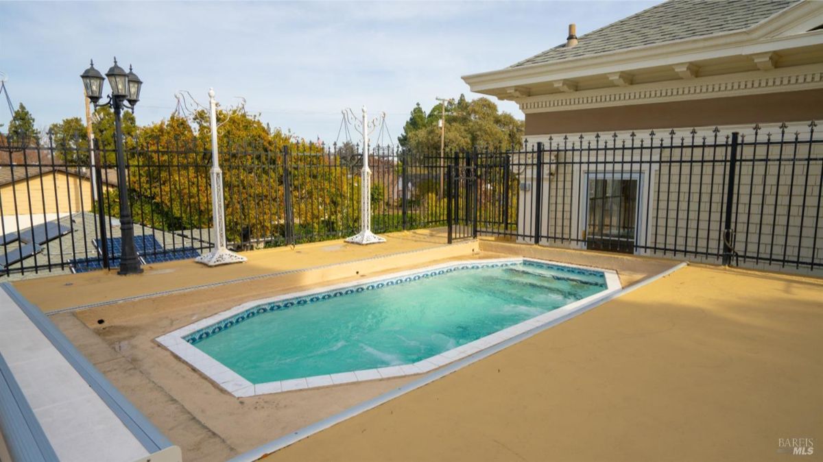 A small, fenced swimming pool located on a rooftop patio with decorative lampposts and open space. The pool is surrounded by a yellowish concrete deck.