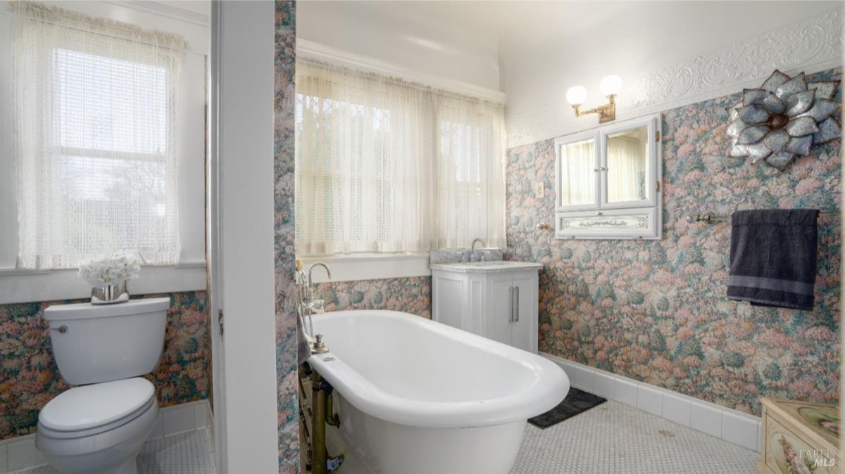 A charming bathroom with floral wallpaper, a clawfoot bathtub, and a window with sheer curtains. The bathroom features vintage-style fixtures and a compact vanity.