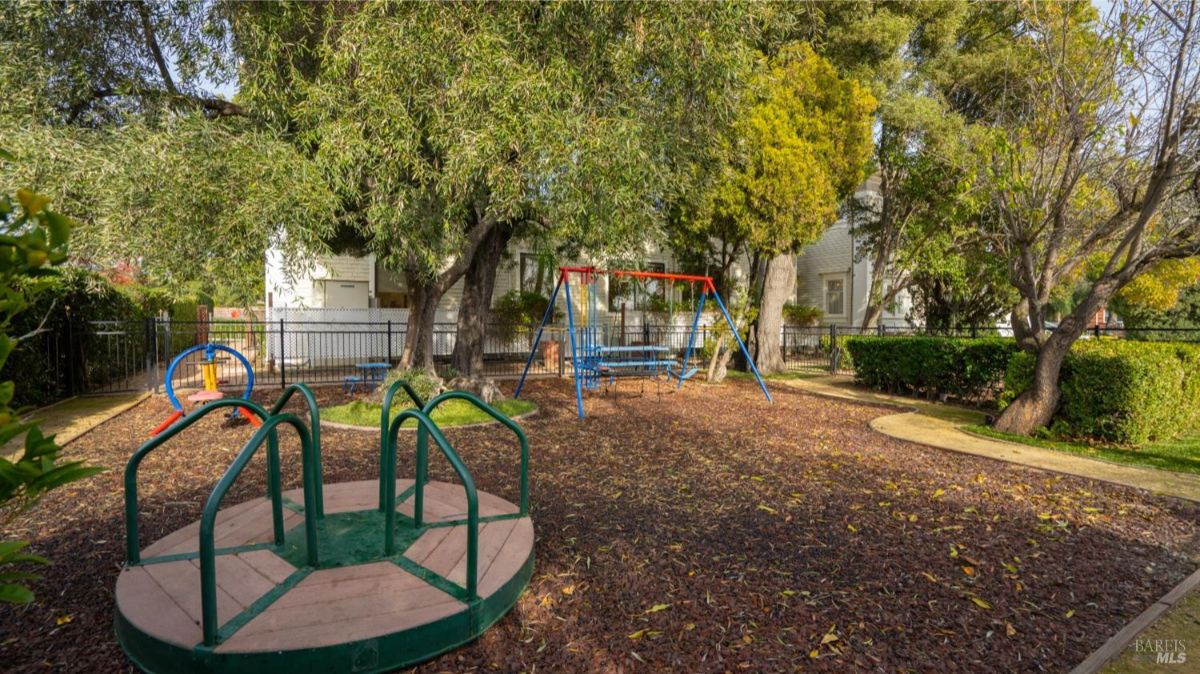 A vibrant playground featuring swings, a merry-go-round, and other play structures. The area is shaded by trees and surrounded by a fence for safety.