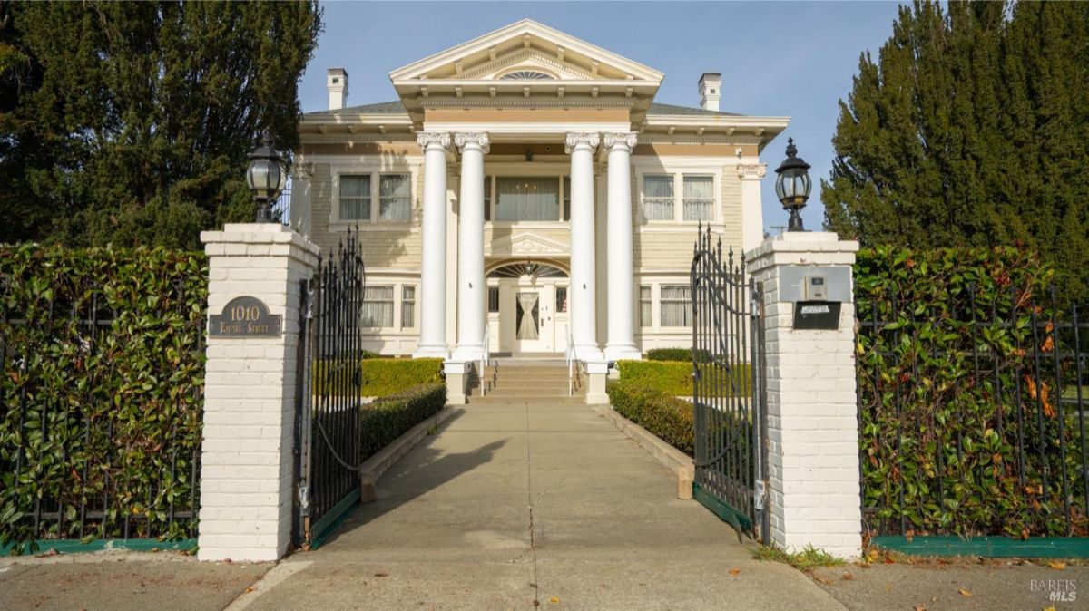 A closer view of the mansion's front entrance, showcasing its prominent white columns and a gated pathway leading to the home. The gates and pillars add an air of exclusivity and elegance to the property.