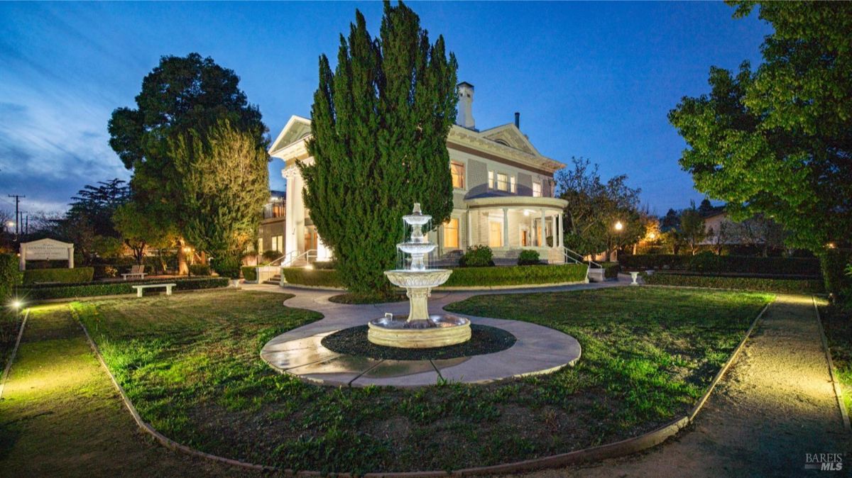 A beautifully illuminated nighttime view of the mansion and its grounds. A central fountain adds charm to the circular pathway, creating an enchanting ambiance around the estate.