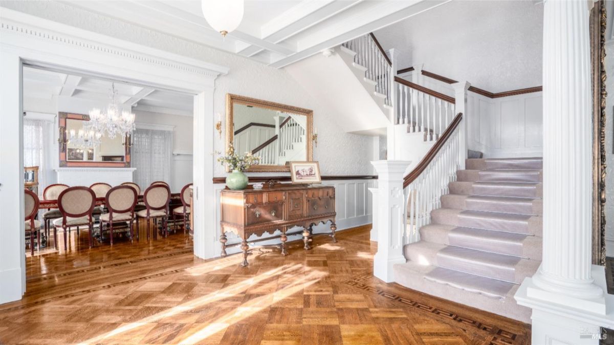  A grand entrance area featuring a staircase with plush carpeting, intricate wood flooring, and white columns. The staircase connects seamlessly to the adjacent formal dining area, visible with its luxurious chandelier and polished wooden furniture.