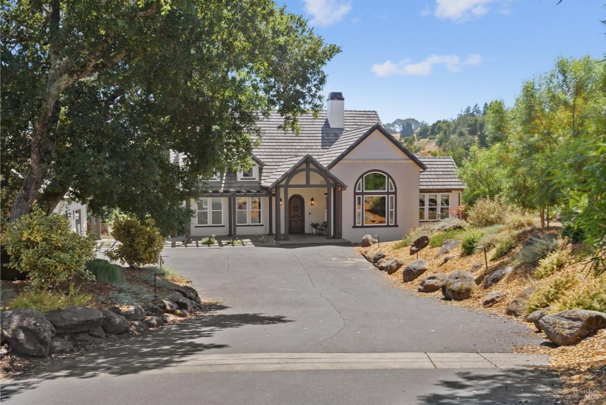 The house has a light-colored exterior, a gabled roof, and a driveway lined with rocks.