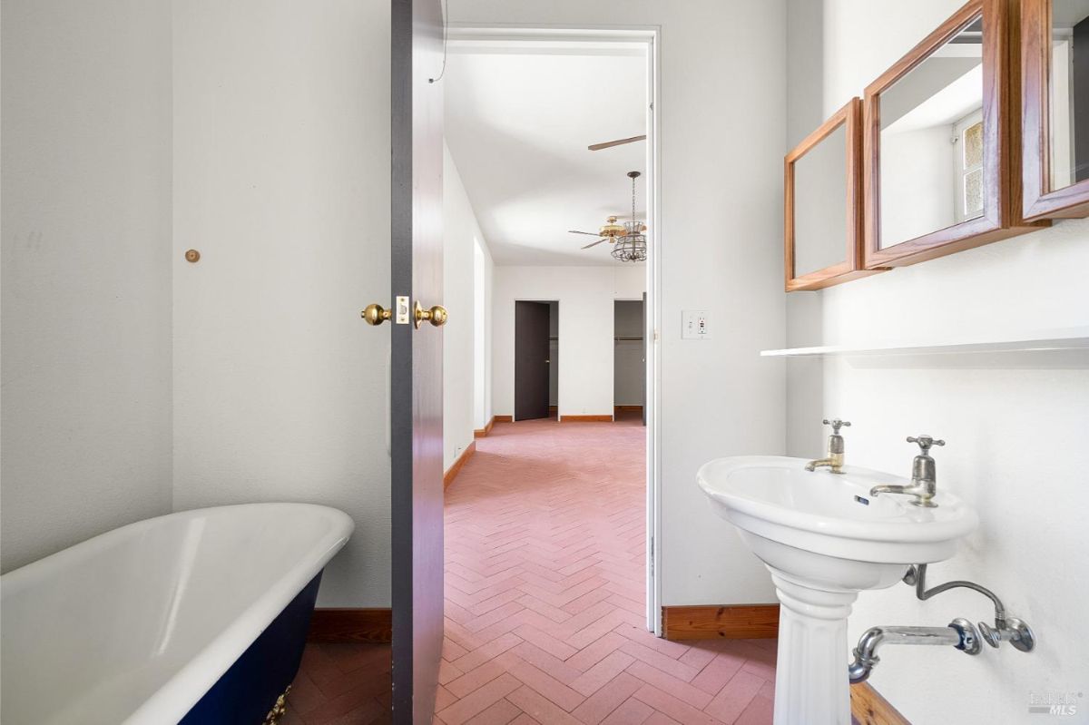 A bathroom with a pedestal sink, a partial view of a clawfoot tub, and a doorway leading to a room with herringbone flooring.