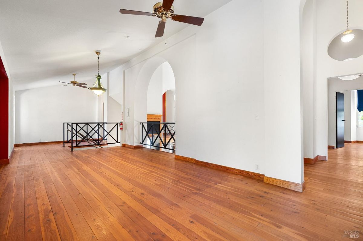 A hallway with wood floors, white walls, ceiling fans, and a railing overlooking a lower area.