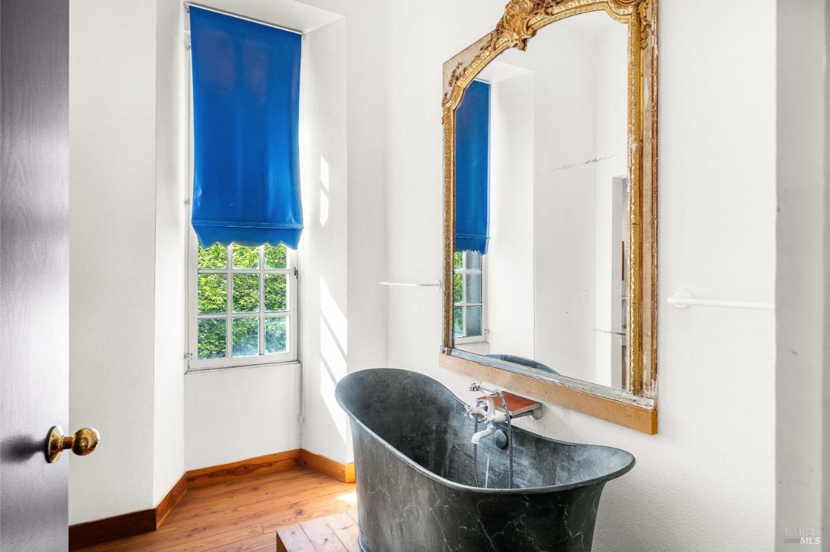 A bathroom with a black freestanding tub, a large ornate mirror, and a window with blue shades.