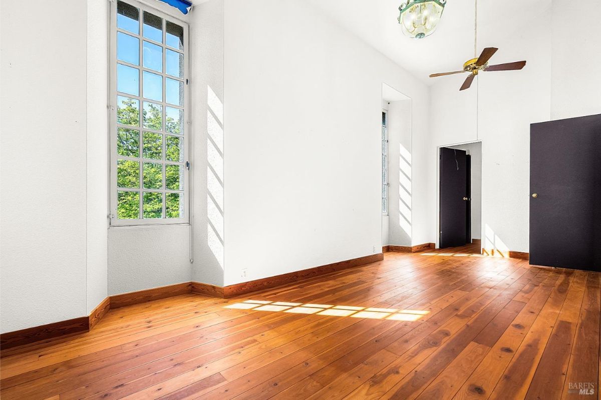 A bright room with tall windows, wood floors, and a ceiling fan under a high ceiling.