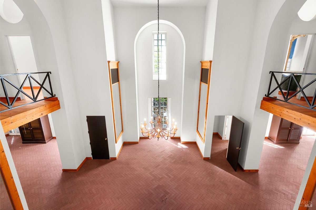 A top-down view of a grand room with a chandelier, tall windows, and upper-level railings.