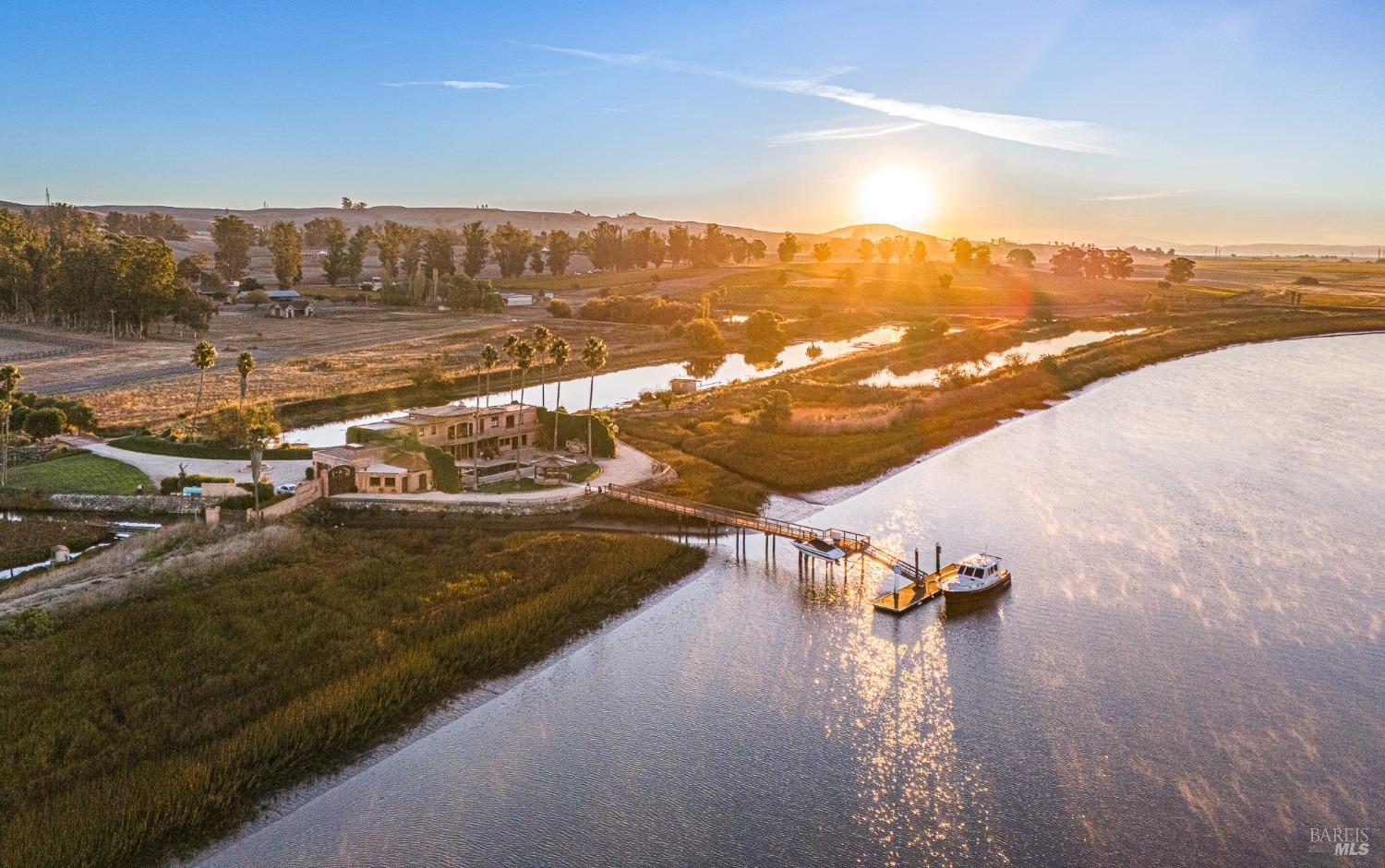 This stunning waterfront estate basks in the golden light of a breathtaking sunrise. Its private dock extends into the calm water, accommodating a boat and smaller watercraft, ready for a day of exploration or relaxation. The surrounding landscape, with its blend of natural beauty and cultivated greenery, provides a tranquil and idyllic setting for this luxurious retreat.