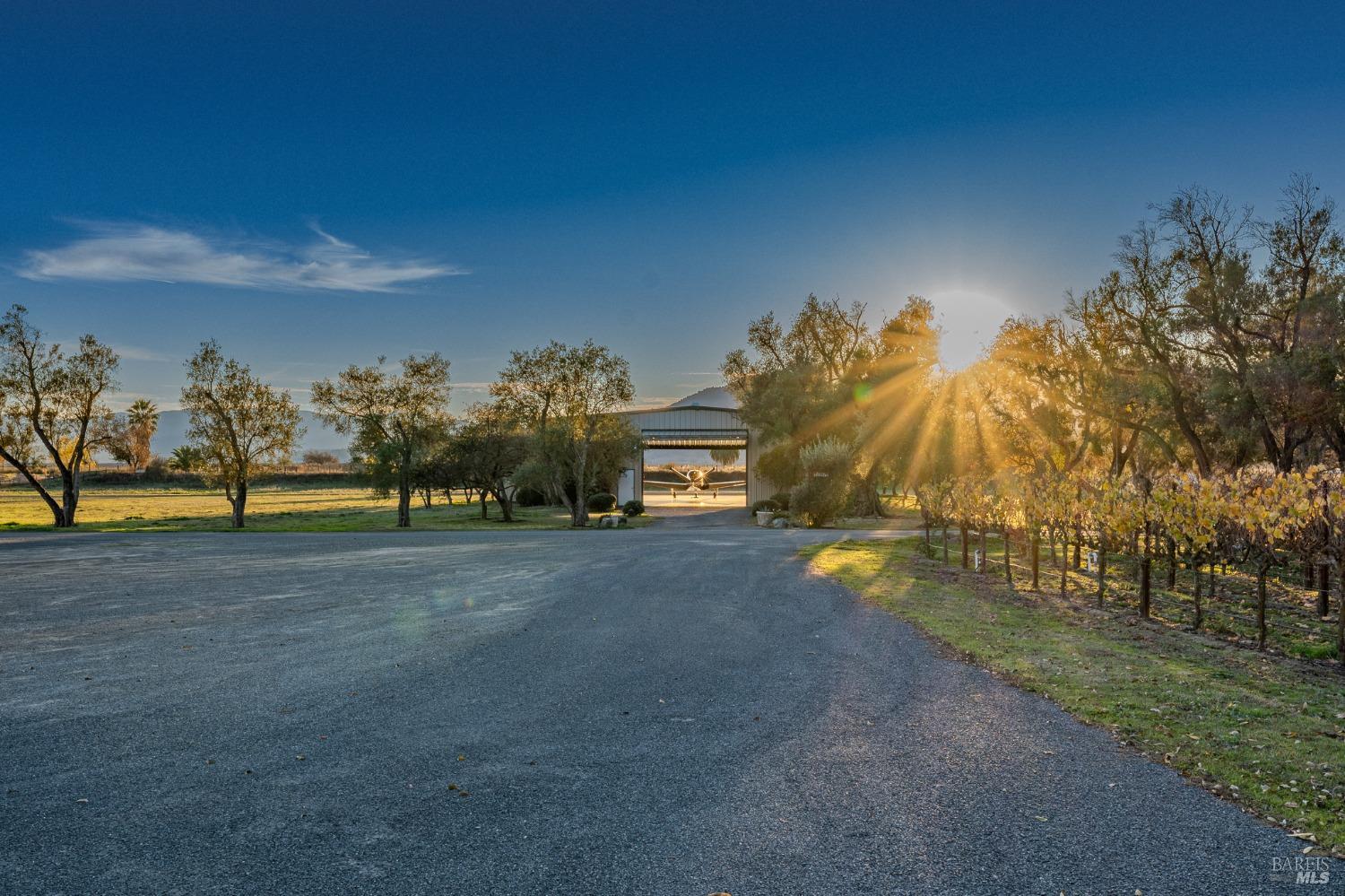 A long driveway, bathed in the warm glow of the setting sun, leads to a large open-sided barn, hinting at the agricultural nature of this expansive property. The surrounding trees and distant mountains create a serene and picturesque rural landscape.