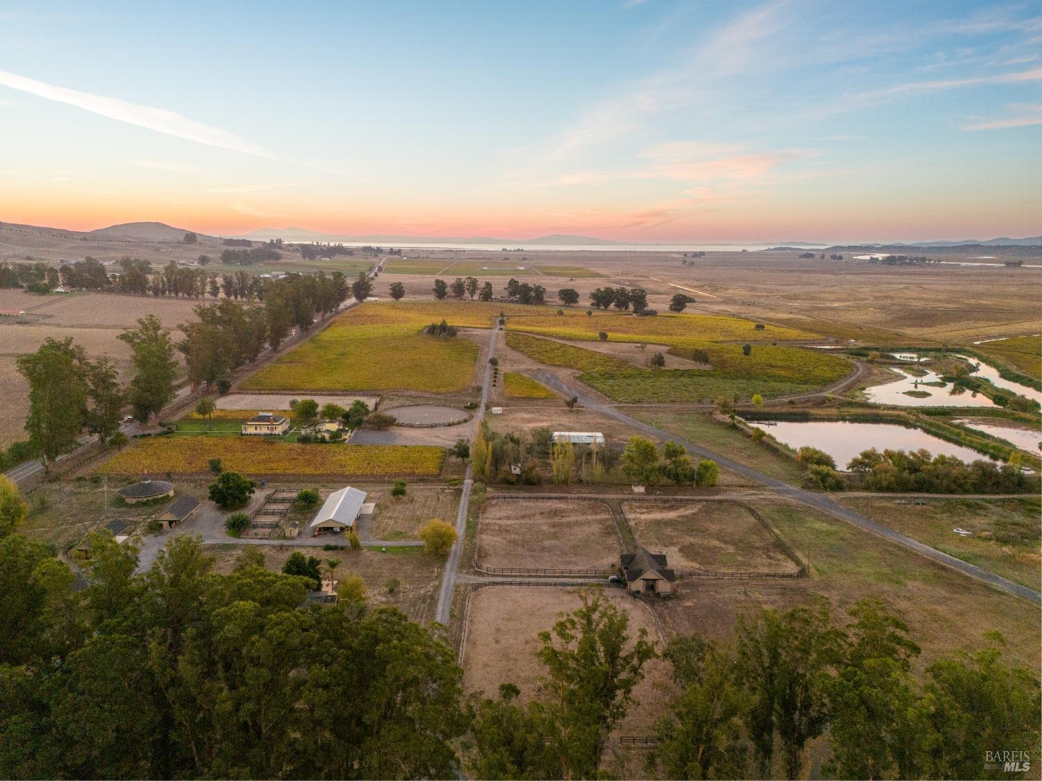 A breathtaking aerial view of a sprawling ranch at sunset. The property features vineyards, paddocks, ponds, and a variety of buildings, all nestled amidst rolling hills and a stunning vista.