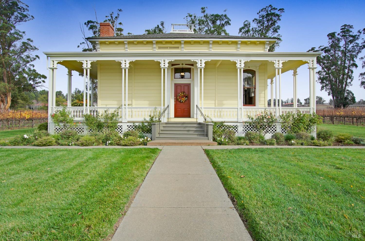 A stately Victorian-era home, painted in a cheerful yellow, features a grand wraparound porch with intricate detailing and a welcoming front entrance. The well-manicured lawn and surrounding greenery enhance the home's classic elegance and curb appeal.