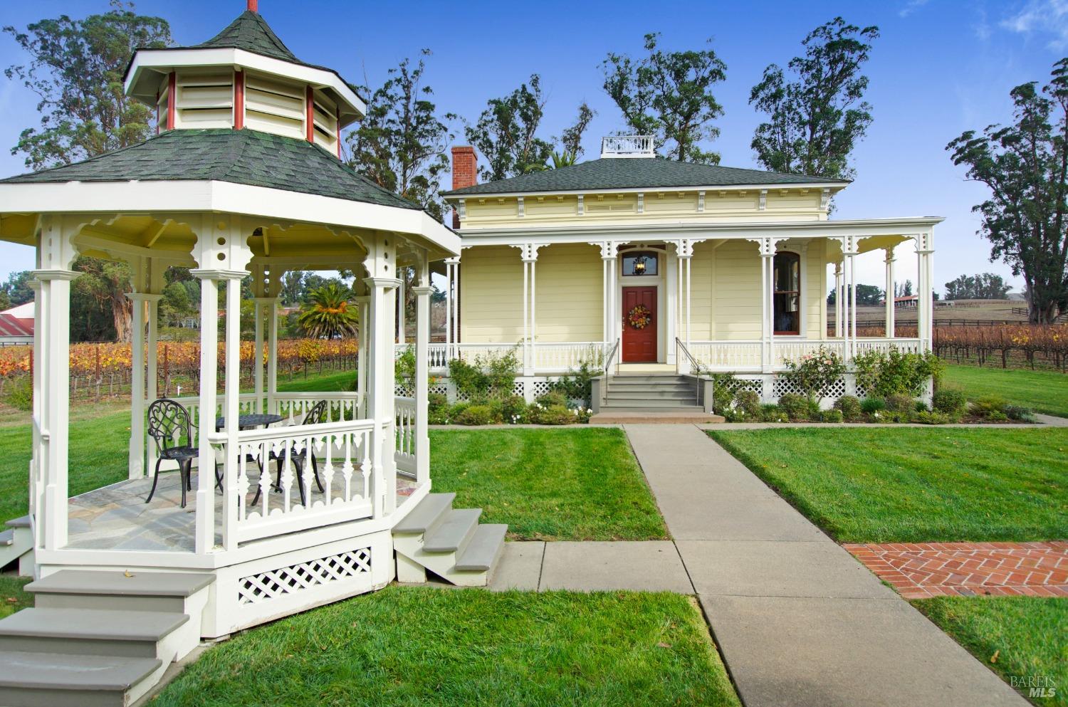 A stately Victorian-era home, painted in a cheerful yellow, features a grand wraparound porch with intricate detailing and a welcoming front entrance. The well-manicured lawn and surrounding greenery enhance the home's classic elegance and curb appeal.