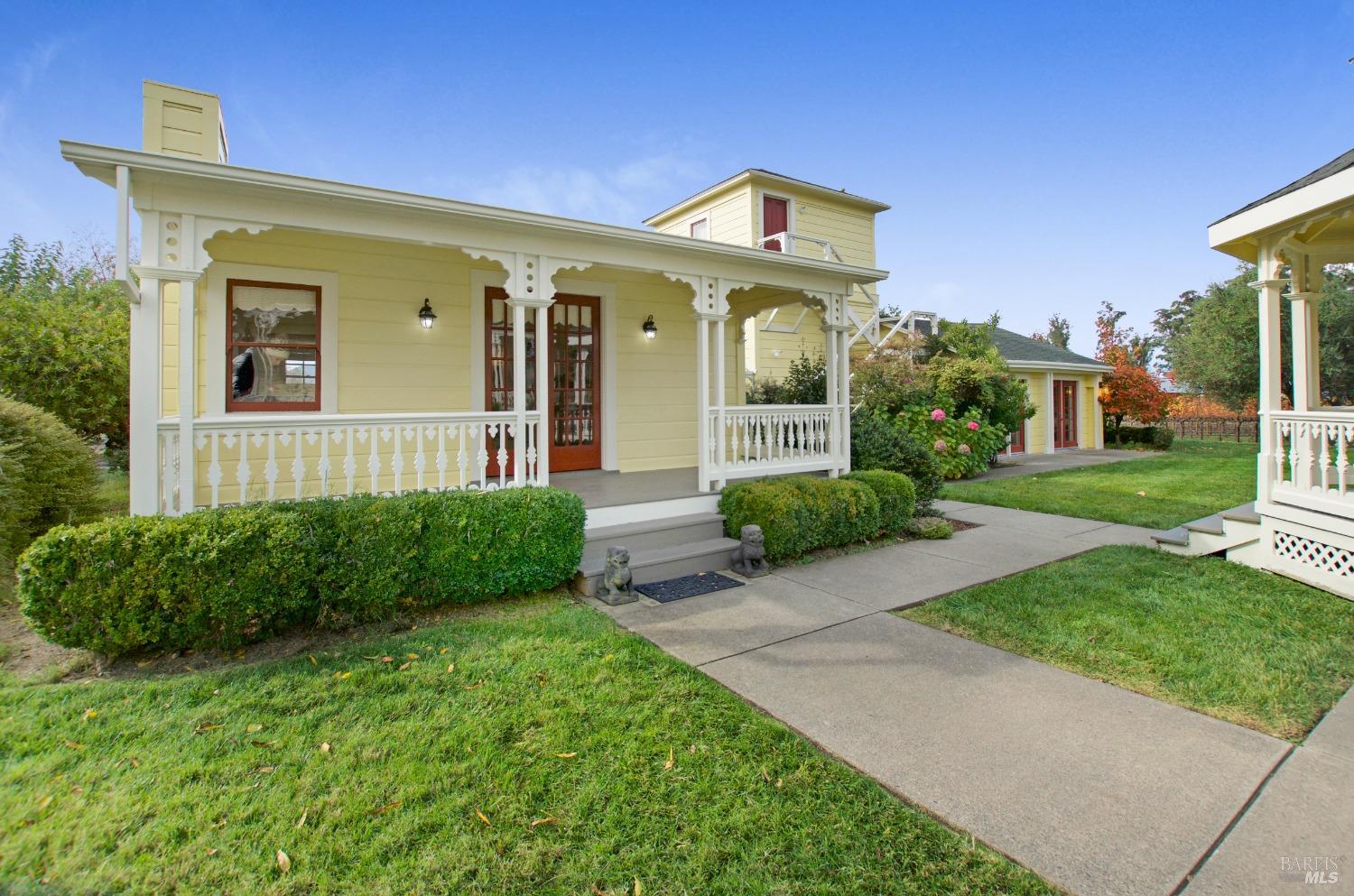 A quaint gazebo and a historic Victorian-style home, painted a cheerful yellow, share a beautifully landscaped lawn, creating a peaceful and inviting scene. The gazebo offers a shaded retreat, while the home's wraparound porch beckons relaxation and enjoyment of the surrounding vineyard views.