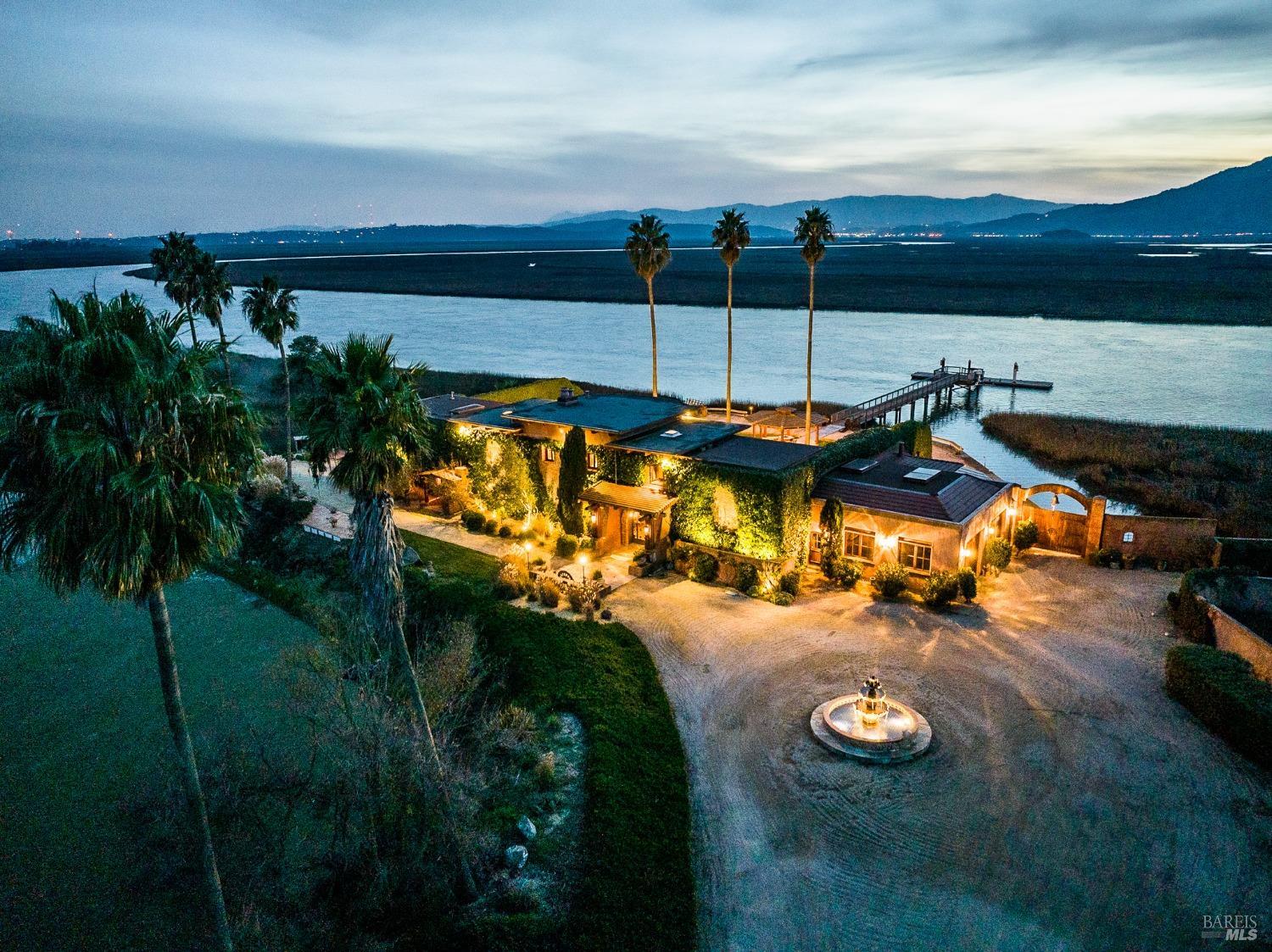 As twilight descends, this luxurious waterfront estate illuminates the night, its warm lights reflecting off the tranquil waters. Palm trees sway gently in the breeze, framing the elegant architecture and creating a truly captivating scene.