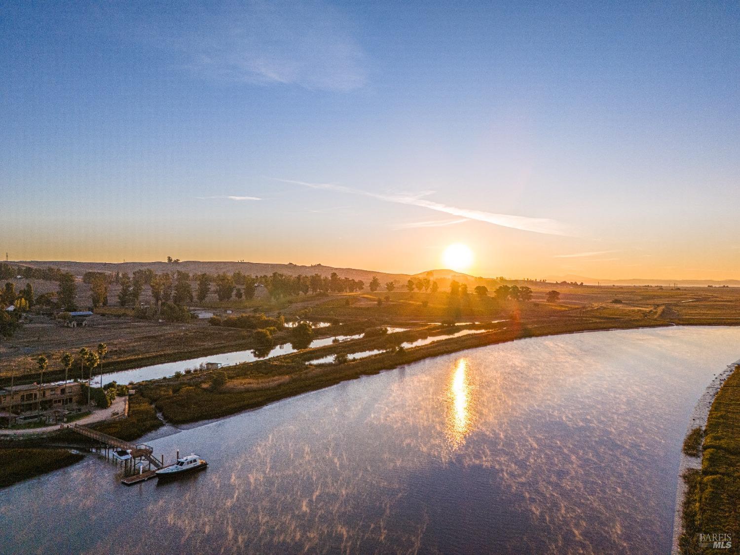 A breathtaking aerial view of a river winding through a serene landscape. The sun is setting, casting a golden glow on the water and the surrounding fields. A small boat is moored to a dock, adding a touch of tranquility to the scene.