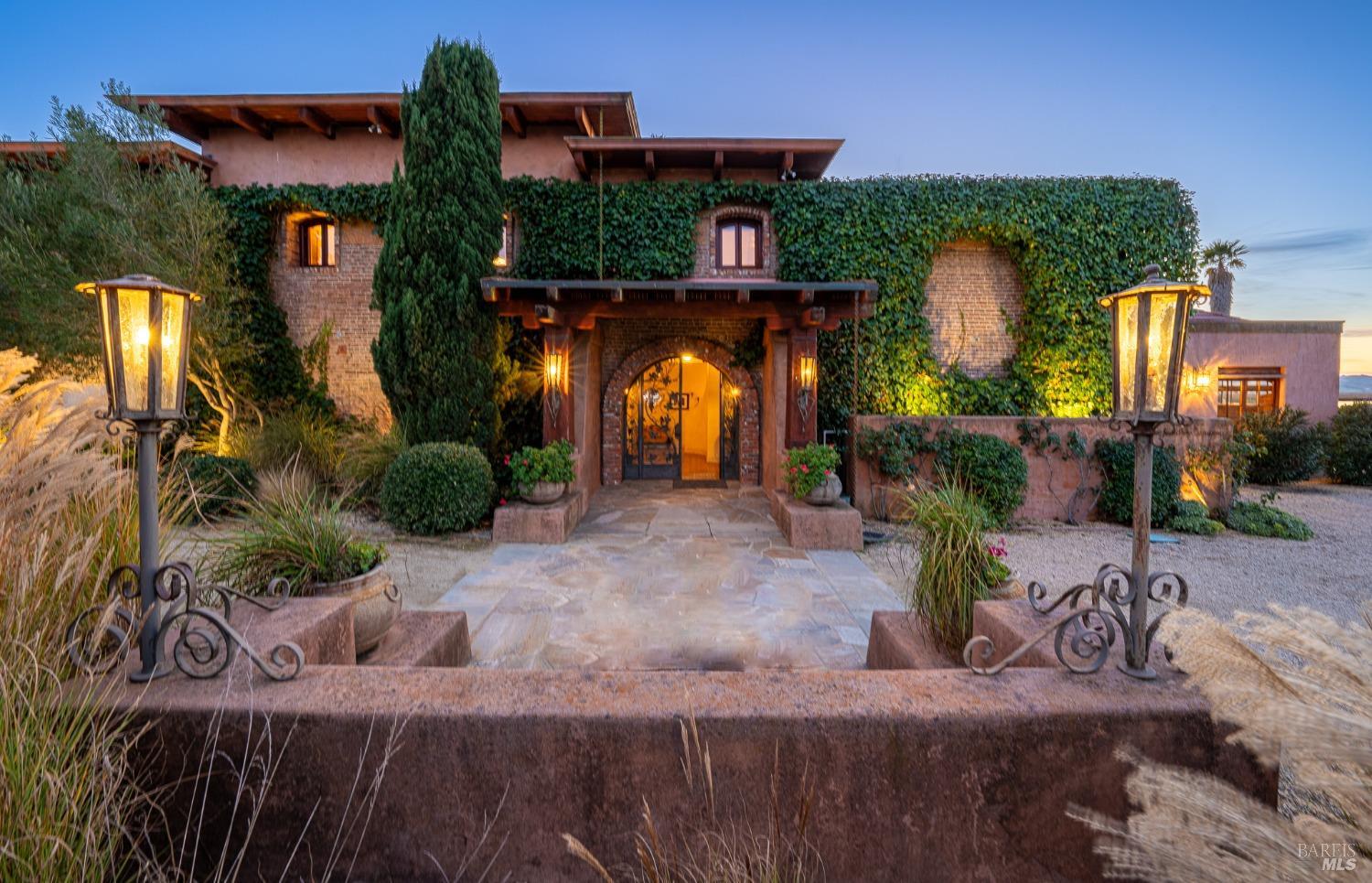 The entrance to this grand estate exudes warmth and elegance, with glowing lanterns illuminating the stone pathway and arched doorway. The ivy-covered walls and surrounding greenery add a touch of rustic charm to this sophisticated residence.