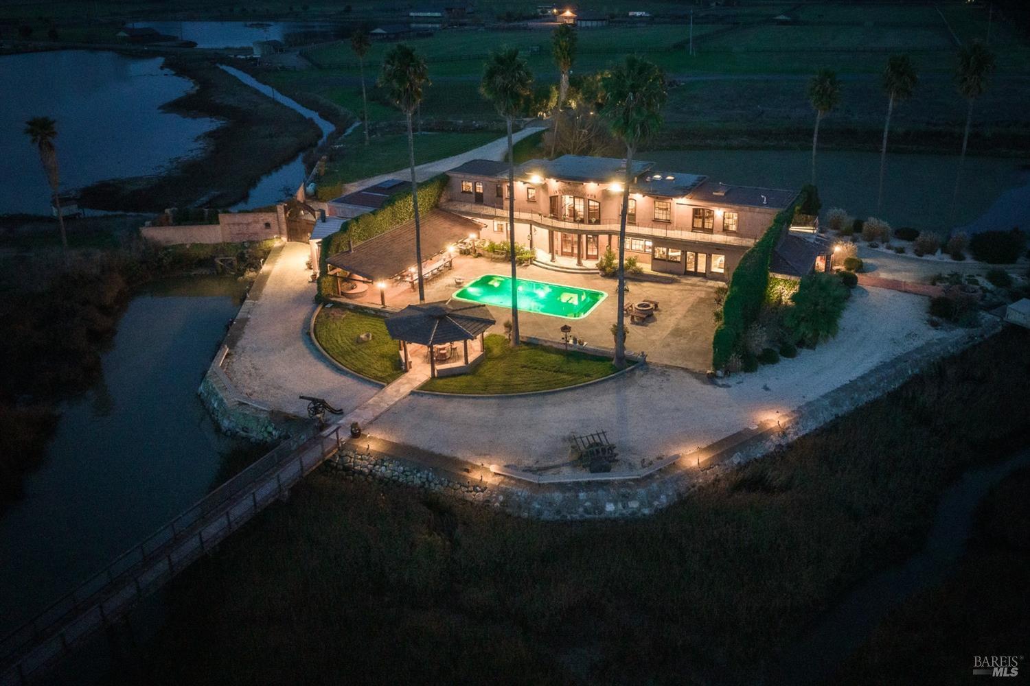 This captivating estate, nestled on its own private island, is a beacon of light against the twilight sky. A pool shimmers under the stars, a gazebo offers a quiet retreat, and a bridge connects the island to the mainland, creating a truly secluded and luxurious haven.