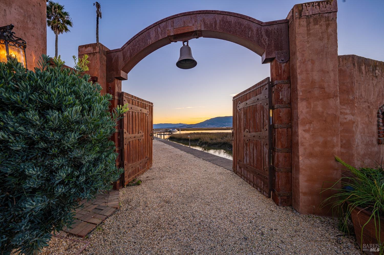 A grand arched gateway, complete with a rustic bell and weathered wooden doors, opens to a picturesque waterfront view, inviting exploration of the estate beyond. The warm hues of the setting sun cast a magical glow on the scene, creating a sense of tranquility and anticipation.