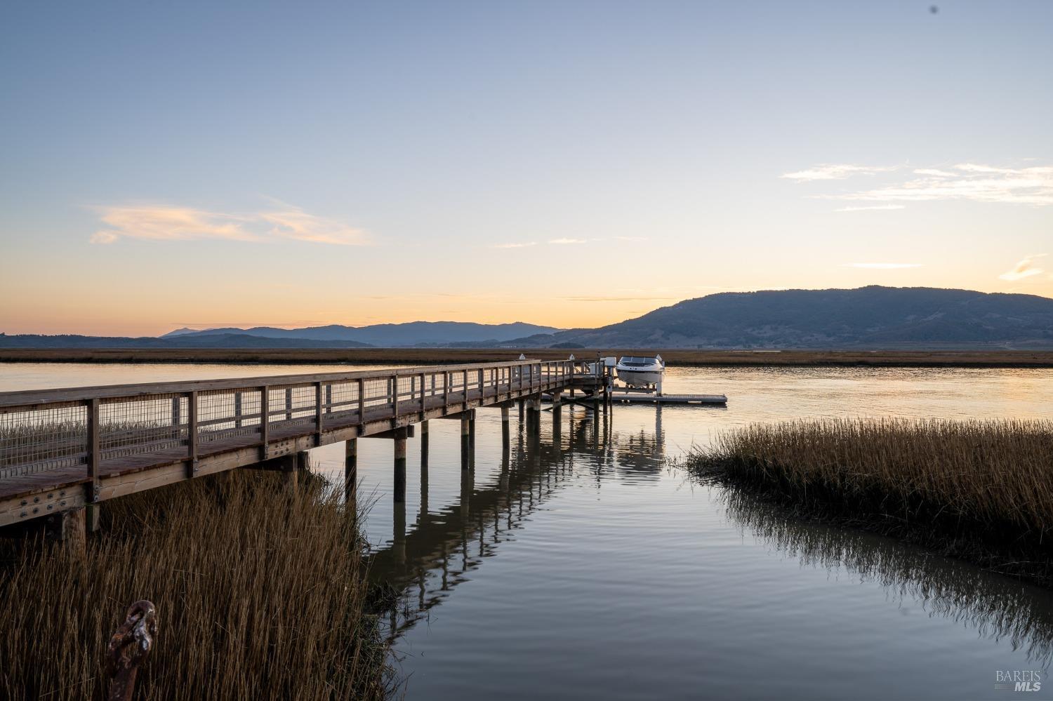 A long wooden pier stretches out into a serene body of water, with a boat gently bobbing at its end, inviting peaceful contemplation as the day draws to a close. The soft hues of the sunset paint the sky, creating a tranquil and picturesque scene.