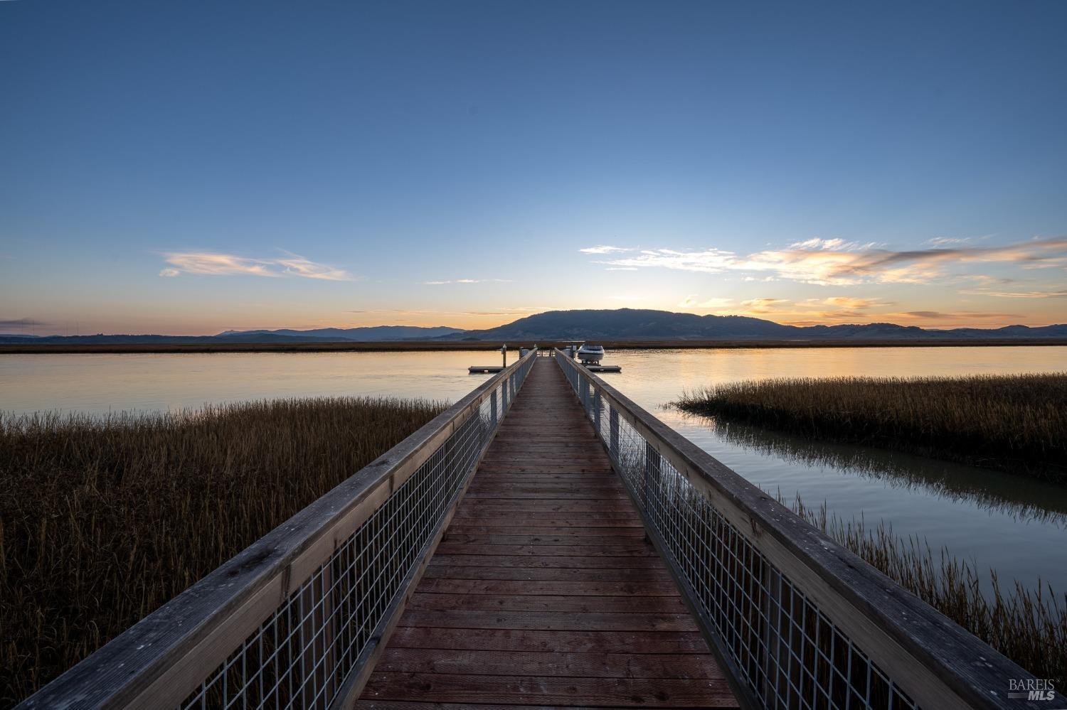 A serene scene of a wooden dock extending into a calm lake at sunset. The sky is painted with hues of orange and purple, reflecting on the still water.