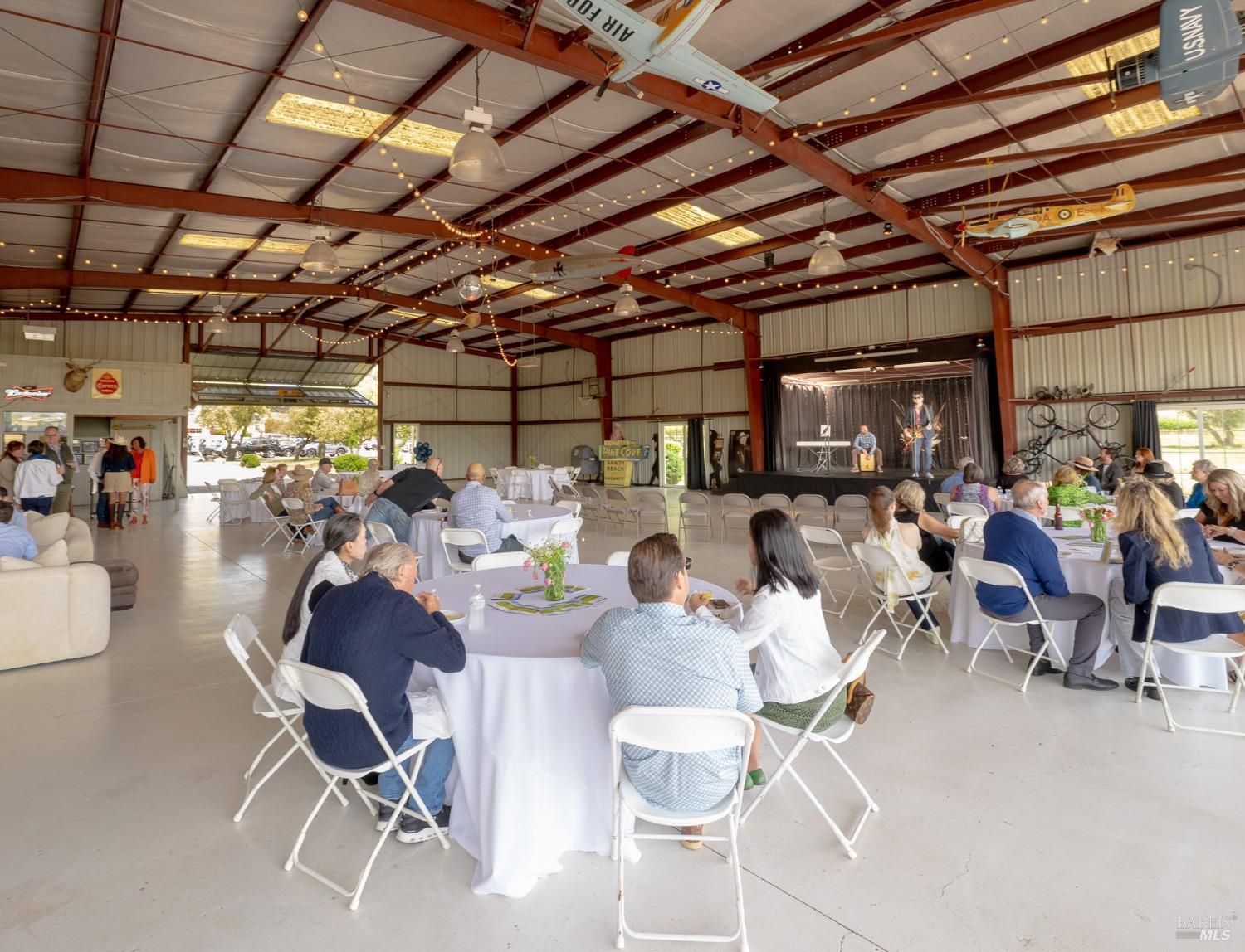 Many people are seated at round tables, and there's a stage at the far end with a few individuals on it, perhaps performing or giving a presentation.