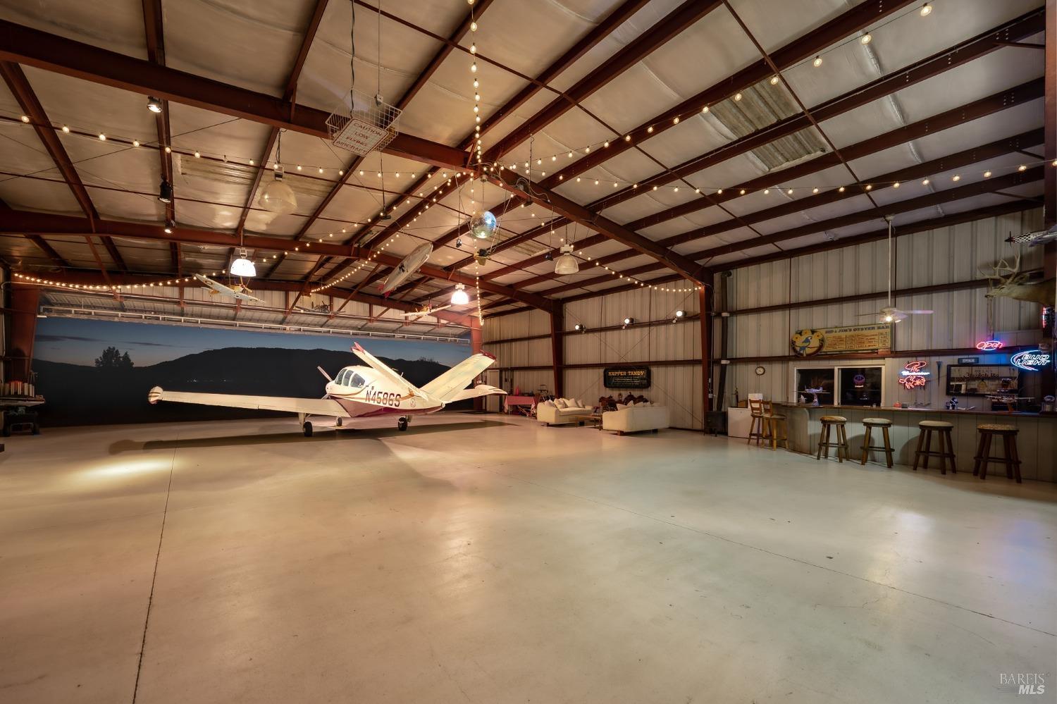 A large, open hangar space with a small plane parked inside. The hangar is well-lit with string lights and features a bar area and seating.